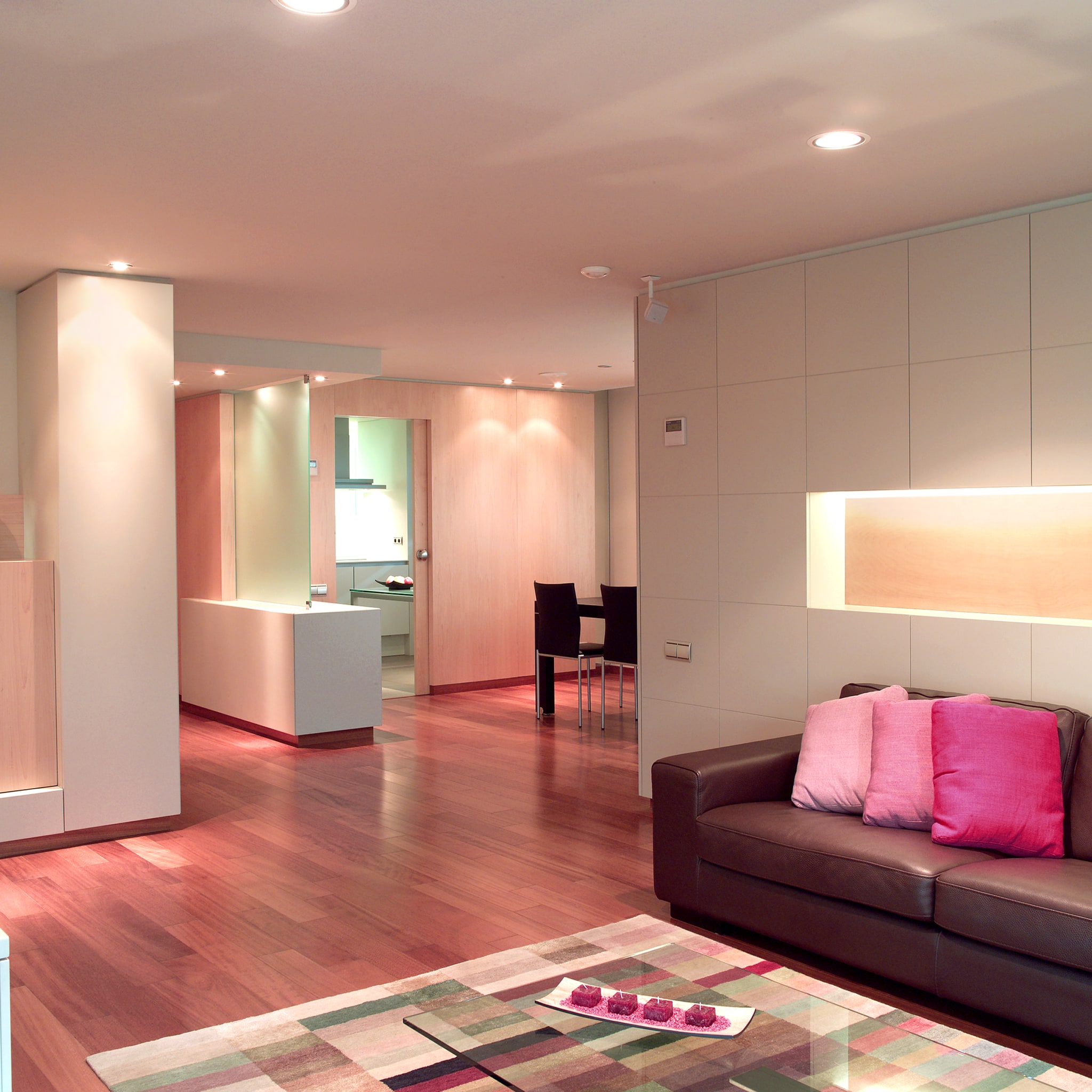 Amplia sala de estar moderna con un cómodo sofá marrón adornado con cojines rosas, suelo de madera cálido y una colorida alfombra geométrica. En el fondo se puede ver un área de comedor minimalista con sillas negras y una cocina elegante.
