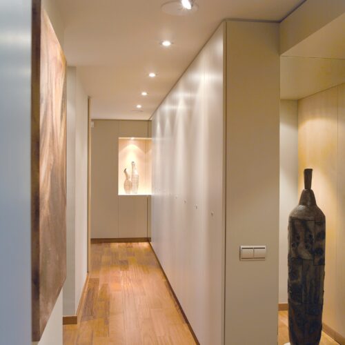 Zonas comunes: Pasillo moderno iluminado que muestra paredes blancas y elegantes, suelos de madera cálida, iluminación empotrada, una alcoba artística con jarrones decorativos, una gran obra de arte abstracto a la izquierda y una figura escultórica de madera a la derecha, creando una atmósfera minimalista y acogedora.
