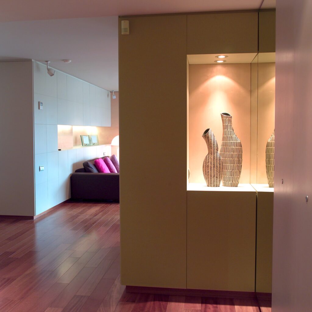 Diseño de interiores moderno en Barcelona: Elegancia y funcionalidad Vista de un moderno espacio de diseño de interiores en Barcelona, con un gabinete amarillo que alberga jarrones decorativos y un sofá oscuro con cojines de colores, resaltando el uso de madera y cristal en su diseño.