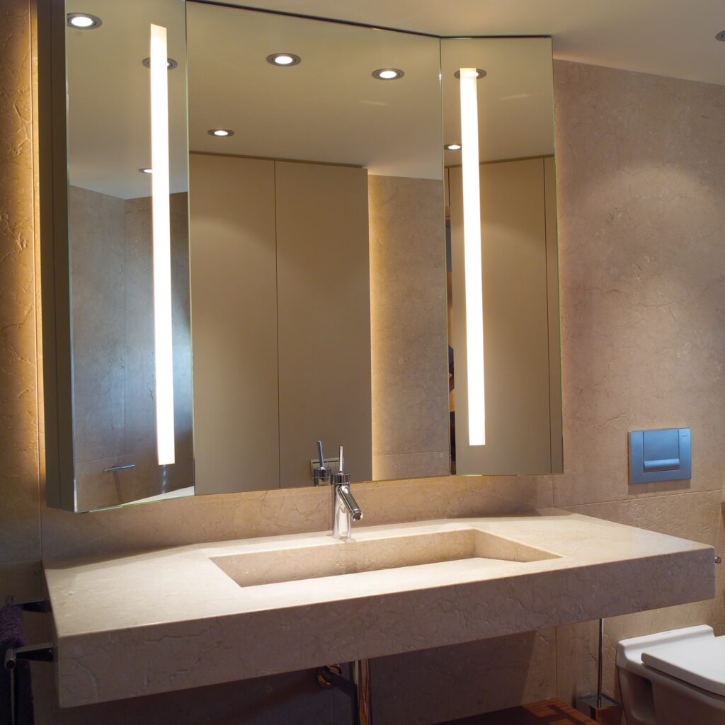 Diseño de baño minimalista moderno con tocador y iluminación elegante Un baño moderno y elegante que muestra un diseño minimalista, con un gran tocador rectangular de superficie de piedra clara, lavabo empotrado, disposición curva de tres paneles espejados, elegantes aparatos de iluminación en el techo y paredes de azulejos en tonos neutros.