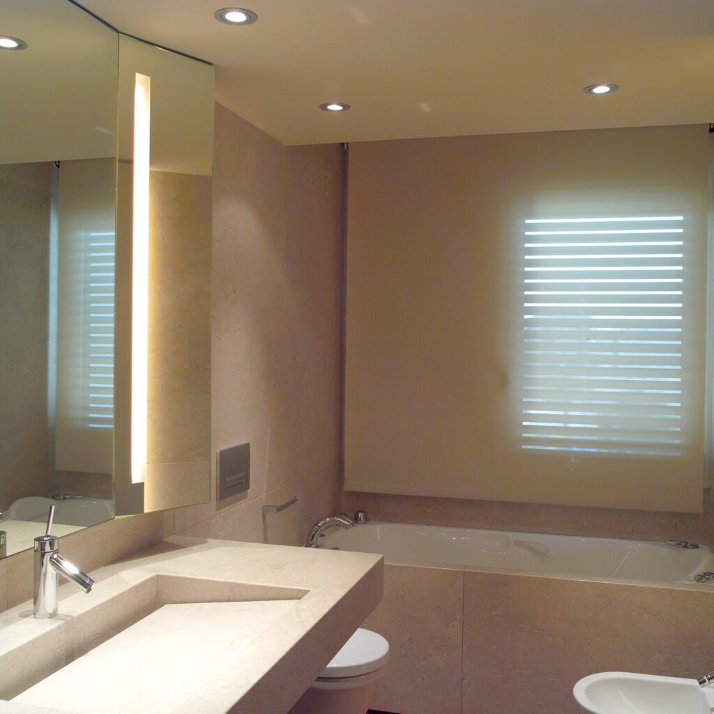 Diseño de interiores moderno y elegante en baño de alta gama en Barcelona Baño moderno y elegante en Barcelona con encimera de piedra, grifo contemporáneo, inodoro de pared minimalista y bañera estilizada, iluminado con luces empotradas y un espejo vertical con luz integrada.