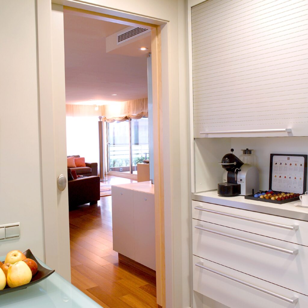 Cocina Moderna con Vista a una Acogedora Sala de Estar y Decoración Funcional Una cocina moderna con una puerta de color claro que conduce a una sala de estar luminosa. En primer plano se muestra un mostrador con un tazón de manzanas variadas, armarios adyacentes con paneles horizontales minimalistas y una cafetera con cápsulas de café. El cálido suelo de madera complementa las grandes ventanas que llenan el espacio de luz natural.
