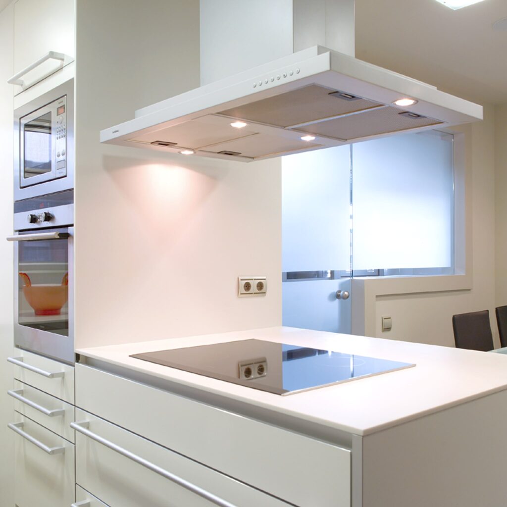 Diseño de cocina moderna y elegante con campana extractora y encimera de vidrio negro Una cocina contemporánea con elegante mobiliario blanco, una encimera de vidrio negro y una campana extractora con iluminación incorporada. El diseño incluye un microondas y un horno empotrados, un tazón naranja brillante dentro del microondas y luz natural iluminando el espacio a través de una puerta de vidrio.