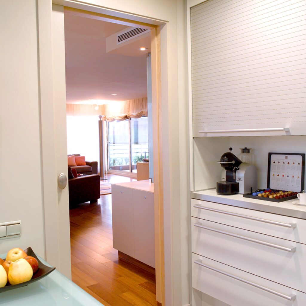 Cocina Brillante y Moderna con Vista a la Acogedora Sala de Estar Una cocina moderna con gabinetes blancos elegantes y una máquina de café espresso, con un plato de manzanas coloridas en primer plano y una vista de una sala de estar acogedora con un sofá marrón y cojines decorativos.