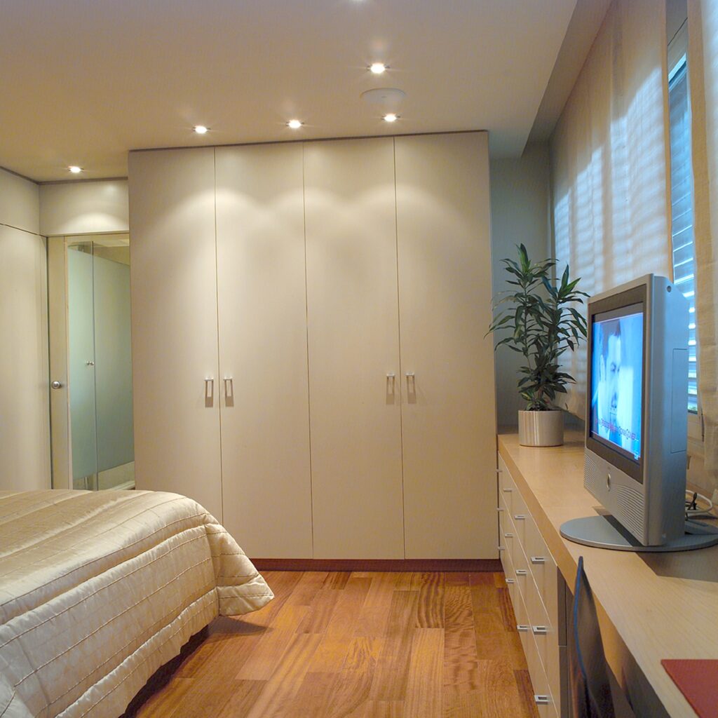 Dormitorio Moderno Acogedor con Estética Natural Un dormitorio acogedor que presenta una paleta de colores claros y cálidos, una cama bien hecha con una manta beige estampada, una cómoda de madera con un televisor, una planta alta de interior en una maceta blanca y un gran espejo que refleja parte de la habitación.