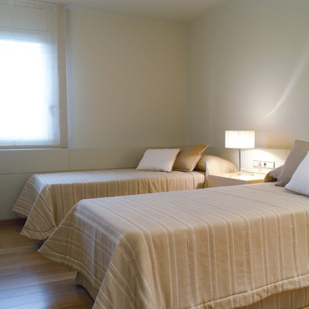 Acogedora Habitación de Invitados con Dos Camas Individuales y Ambiente Cálido Una acogedora habitación de invitados con dos camas individuales con ropa de cama de rayas beige claro, una mesita de noche con una lámpara moderna y cortinas suaves que permiten la entrada de luz natural. La decoración minimalista crea una atmósfera cálida y acogedora, perfecta para relajarse o pasar la noche.