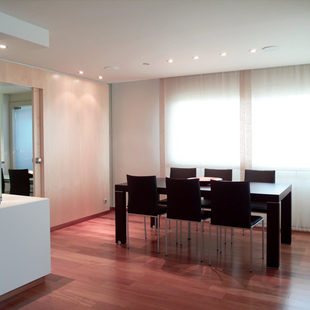 Área de Comedor Moderna con Mesa de Madera Elegante y Sillas Negras Un área de comedor contemporánea con una mesa de madera estilizada rodeada por seis sillas negras, con un piso de madera dura y luces empotradas en el techo. Cortinas grandes y translúcidas permiten que la suave luz natural ilumine el espacio, realzando la atmósfera acogedora.