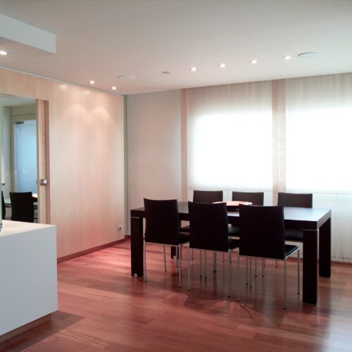 Un área de comedor contemporánea con una mesa de madera estilizada rodeada por seis sillas negras, con un piso de madera dura y luces empotradas en el techo. Cortinas grandes y translúcidas permiten que la suave luz natural ilumine el espacio, realzando la atmósfera acogedora.