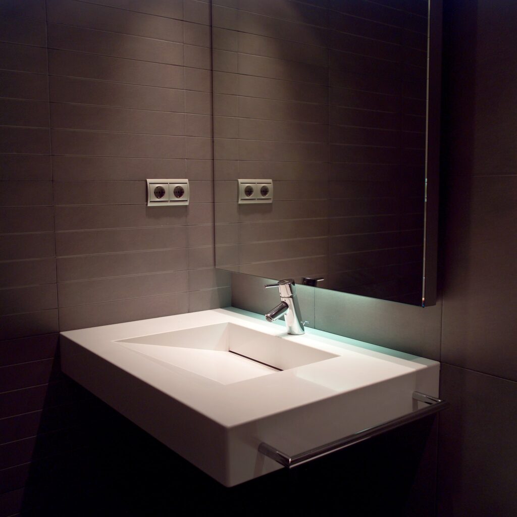 Diseño de lavabo de baño moderno y elegante con estética minimalista Un lavabo de baño moderno y cuadrado en blanco, con un elegante grifo plateado. El lavabo está ubicado contra paredes de azulejos gris oscuro e incluye un gran espejo que refleja el entorno, con tomas de corriente visibles arriba. El espacio se realza con una suave iluminación ambiental.
