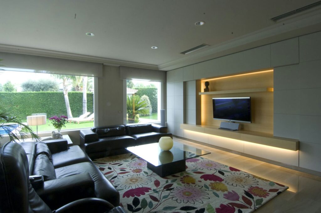 Moderno salón de diseño de interiores en Barcelona con sofá de cuero y mesa de vidrio Salón contemporáneo en Barcelona que destaca por su elegante sofá de cuero negro y mesa de cristal, iluminado de forma natural. Ventanas amplias ofrecen vistas de un jardín exterior, resaltando la funcionalidad y estética del diseño de alta gama.