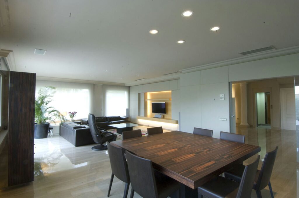 Salón y Comedor Moderno de Alta Gama en Barcelona Amplio y moderno espacio de salón y comedor en Barcelona, con mesa de madera oscura, elegantes sillas, un sofá negro y acabados de mármol en el suelo, iluminado por luz natural a través de ventanas amplias.