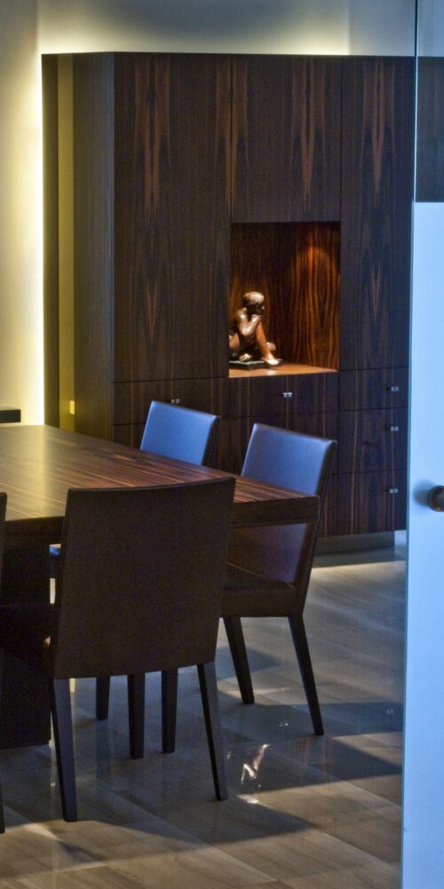 Moderno comedor en Barcelona con mesa de madera y sillas tapizadas oscuras, acompañado de un armario de madera con iluminación de nicho. Diseño funcional y estético, con materiales de alta calidad en un ambiente contemporáneo.