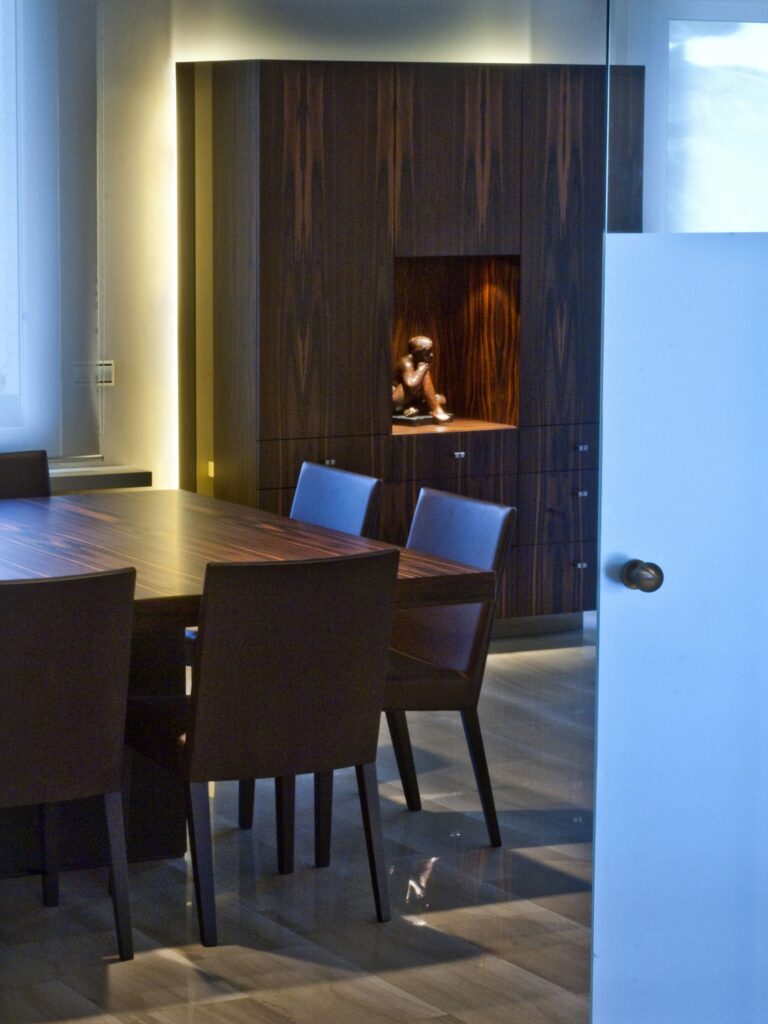 Diseño de interiores moderno en Barcelona: Comedor elegante Moderno comedor en Barcelona con mesa de madera y sillas tapizadas oscuras, acompañado de un armario de madera con iluminación de nicho. Diseño funcional y estético, con materiales de alta calidad en un ambiente contemporáneo.