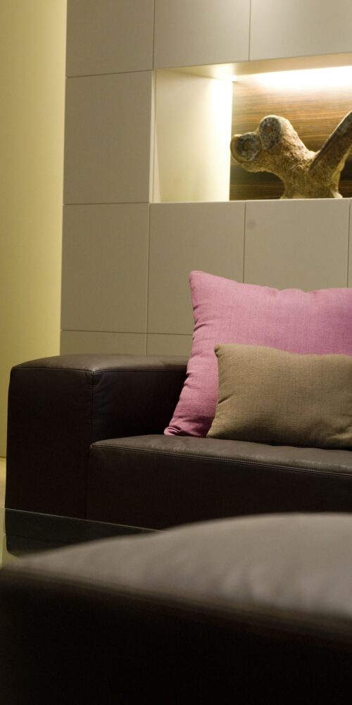 Una sala de estar moderna con un sofá de cuero oscuro, cojines decorativos en tonos rosa y beige, diseño de pared minimalista con iluminación integrada y una mesa de café de vidrio con una superficie decorada.