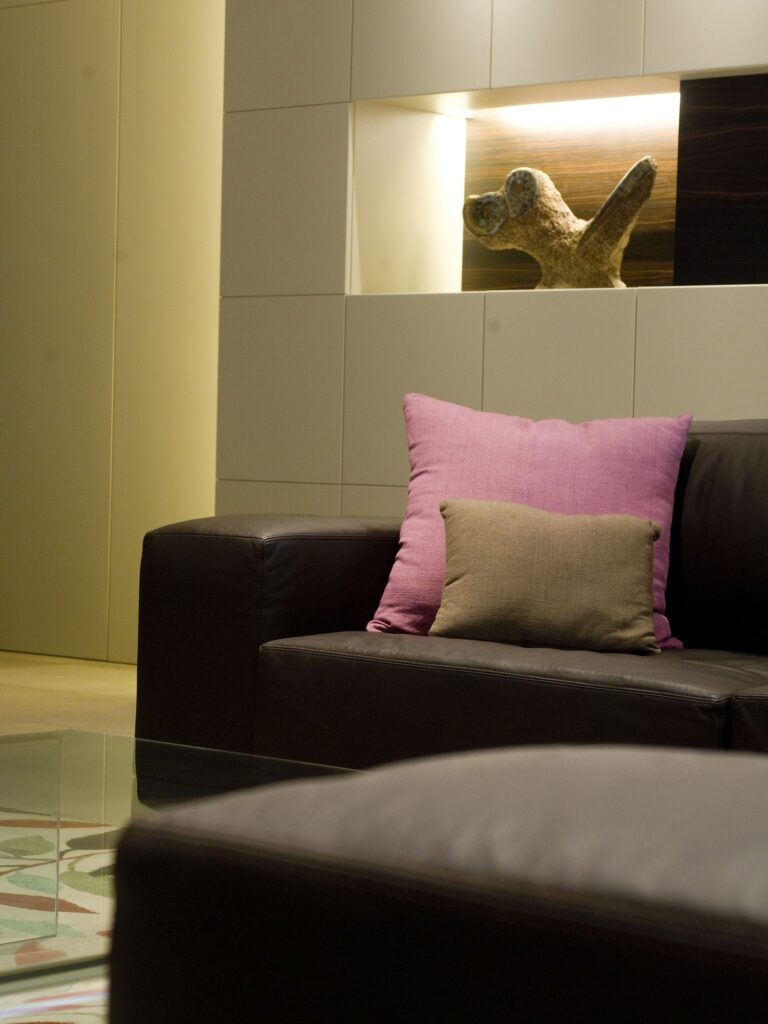 Sala de estar moderna y acogedora con sofá de cuero oscuro y diseño minimalista Una sala de estar moderna con un sofá de cuero oscuro, cojines decorativos en tonos rosa y beige, diseño de pared minimalista con iluminación integrada y una mesa de café de vidrio con una superficie decorada.