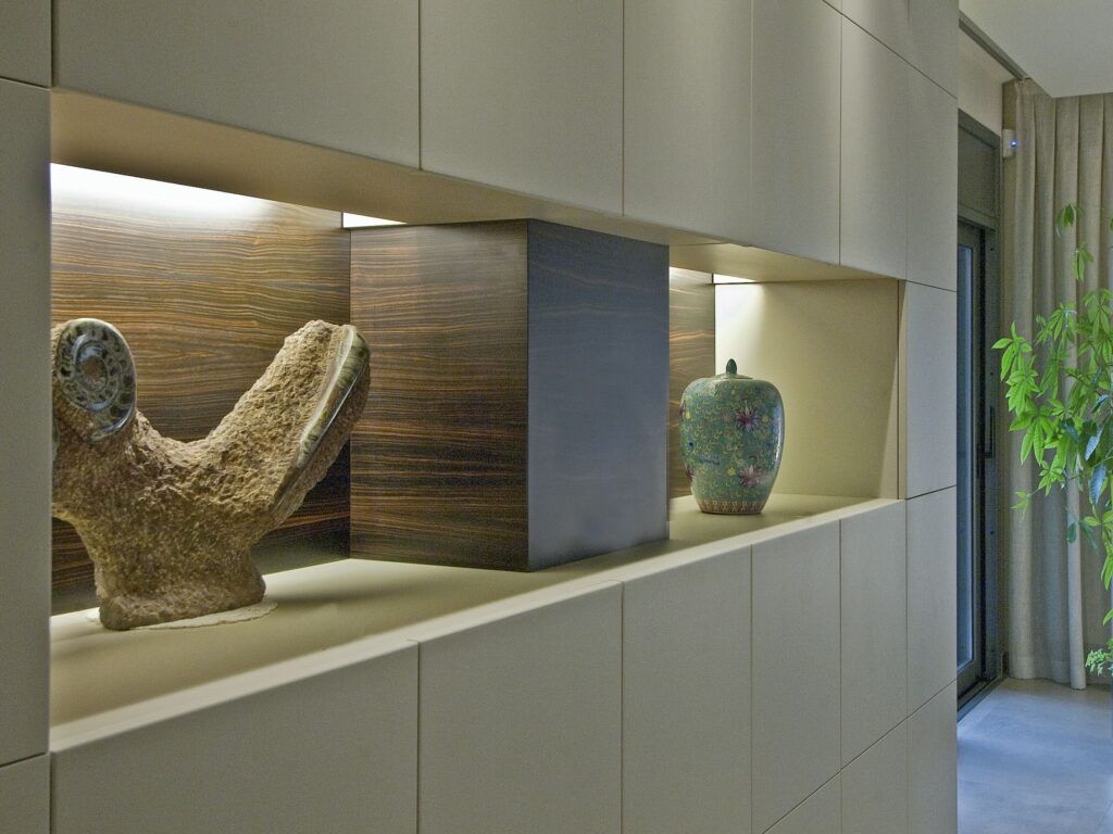 Acentos de Diseño Interior Contemporáneo con Exhibiciones Artísticas Un ambiente interior moderno que presenta una estantería elegante con tres elementos únicos: una escultura texturada, una caja rectangular oscura y un jarrón verde decorativo con patrones florales, todos acentuados por una iluminación suave y un fondo de paneles de pared neutros.