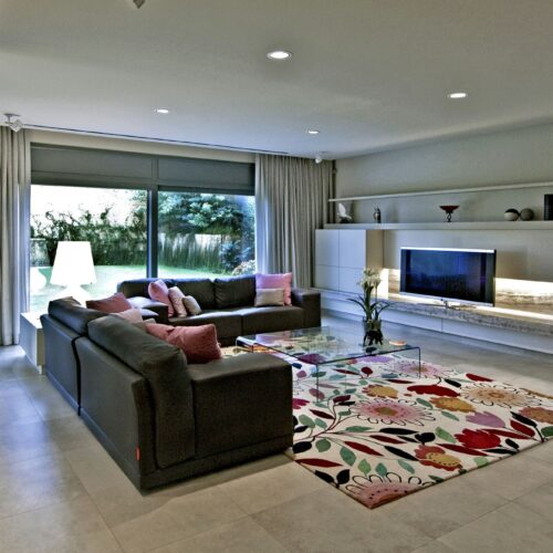 Casa en Calle Llevant: Un amplio interior de salón moderno con un sofá seccional oscuro y cojines de colores, una mesa de café de cristal, una alfombra de diseño floral vibrante, una unidad de entretenimiento minimalista con un televisor de pantalla plana, plantas en macetas y cortinas ligeras que enmarcan grandes ventanas que permiten que la luz natural inunde el espacio.