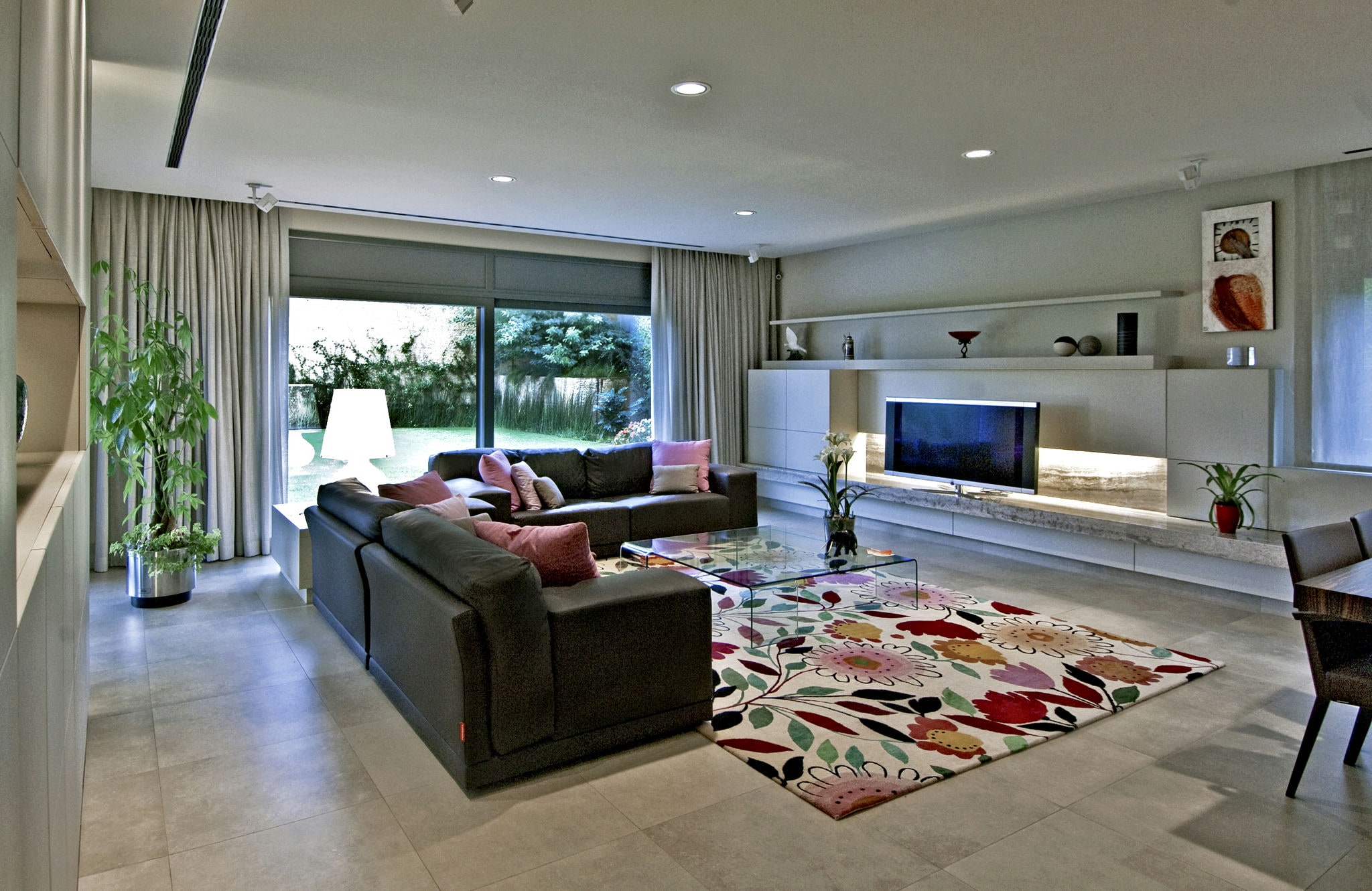 Casa en Calle Llevant: Un amplio interior de salón moderno con un sofá seccional oscuro y cojines de colores, una mesa de café de cristal, una alfombra de diseño floral vibrante, una unidad de entretenimiento minimalista con un televisor de pantalla plana, plantas en macetas y cortinas ligeras que enmarcan grandes ventanas que permiten que la luz natural inunde el espacio.