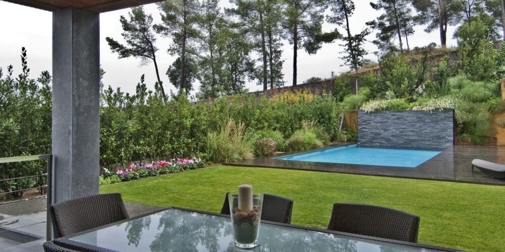 Un jardín moderno al aire libre que presenta una mesa de comedor con tapa de cristal y sillas de mimbre negro, rodeado de exuberantes céspedes verdes y vibrantes plantas florecientes. En el fondo, una elegante piscina rectangular está enmarcada por paredes de piedra y varios árboles, todo bajo un cielo nublado, creando una atmósfera tranquila para la relajación.