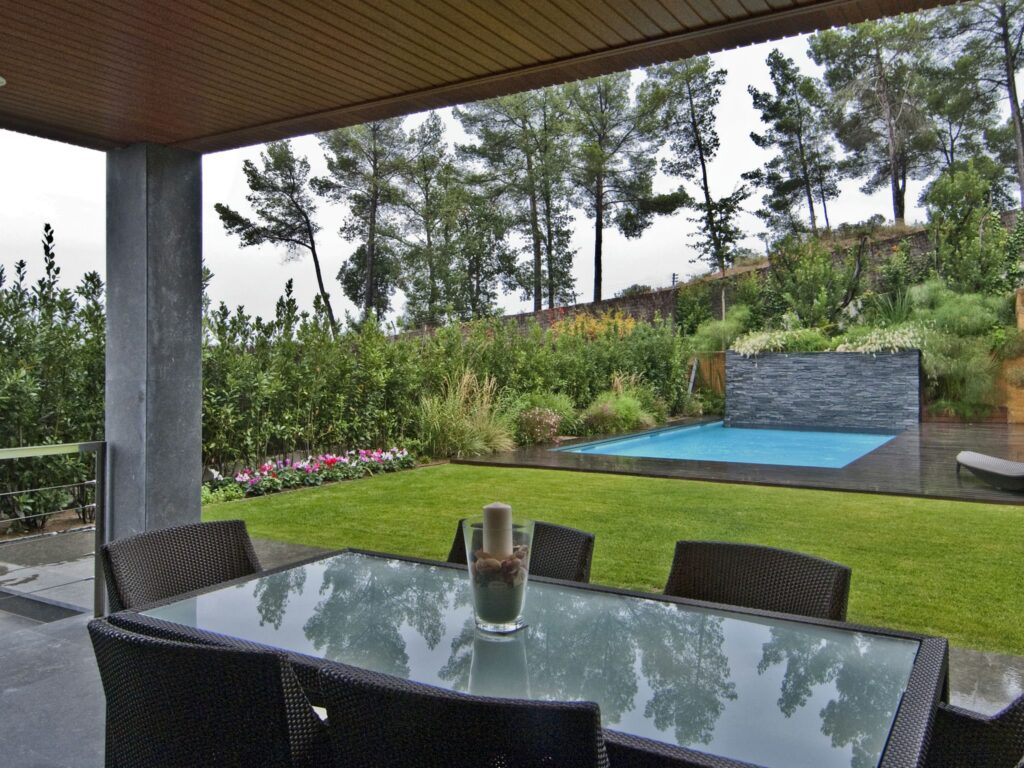 Sereno Jardín Moderno al Aire Libre con Mesa de Comedor de Cristal y Piscina Un jardín moderno al aire libre que presenta una mesa de comedor con tapa de cristal y sillas de mimbre negro, rodeado de exuberantes céspedes verdes y vibrantes plantas florecientes. En el fondo, una elegante piscina rectangular está enmarcada por paredes de piedra y varios árboles, todo bajo un cielo nublado, creando una atmósfera tranquila para la relajación.