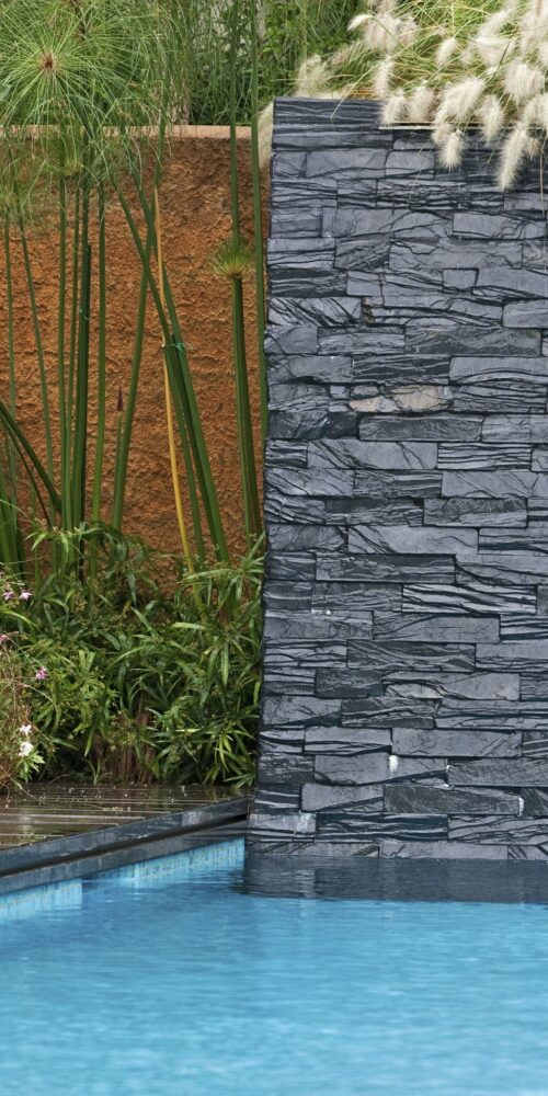 Una escena serena al aire libre que presenta una piscina moderna con agua azul cristalina, bordeada por un decking de piedra oscura y vegetación exuberante, incluyendo plantas altas y flores coloridas, complementada por una pared vertical de piedra contemporánea y un fondo texturado de color naranja.