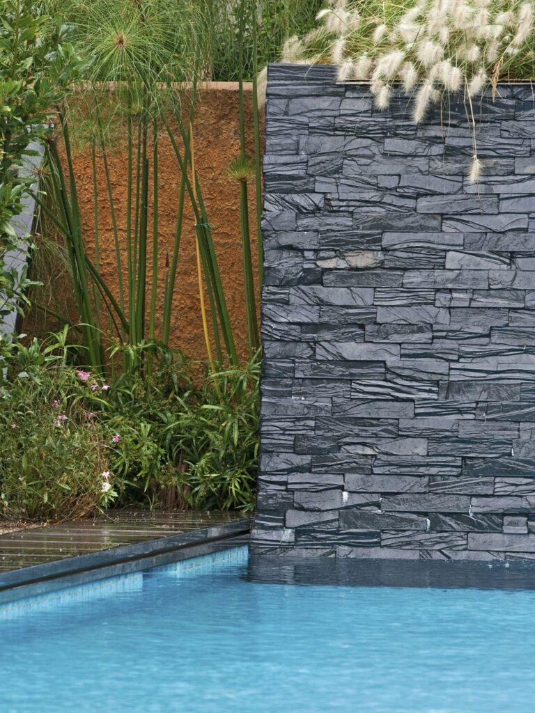 Serenidad Moderna: Una Tranquila Piscina Rodeada de Vegetación Exuberante Una escena serena al aire libre que presenta una piscina moderna con agua azul cristalina, bordeada por un decking de piedra oscura y vegetación exuberante, incluyendo plantas altas y flores coloridas, complementada por una pared vertical de piedra contemporánea y un fondo texturado de color naranja.