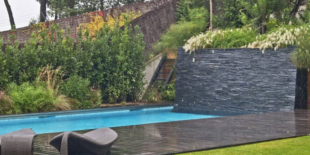 Una escena exterior tranquila que presenta una piscina rectangular con agua clara y azul, dos elegantes tumbonas curvadas en una terraza de madera, y una pared vertical de piedra adornada con vegetación, rodeada de plantas y árboles vibrantes.