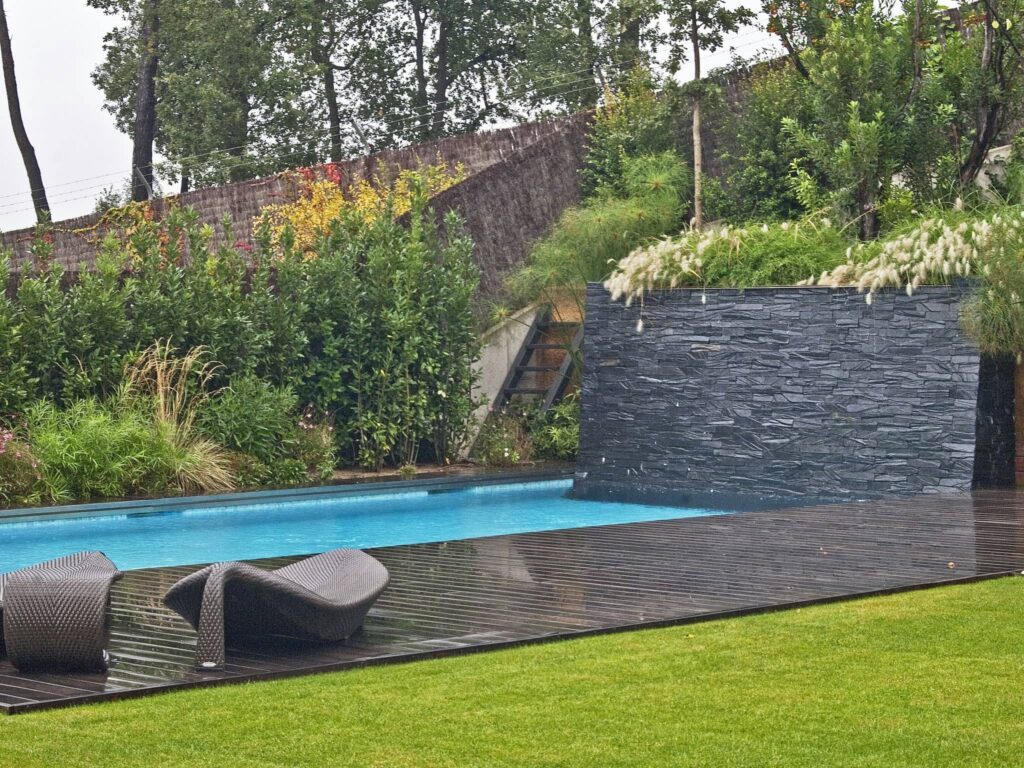 Serena piscina al aire libre con tumbonas curvadas y vegetación exuberante Una escena exterior tranquila que presenta una piscina rectangular con agua clara y azul, dos elegantes tumbonas curvadas en una terraza de madera, y una pared vertical de piedra adornada con vegetación, rodeada de plantas y árboles vibrantes.