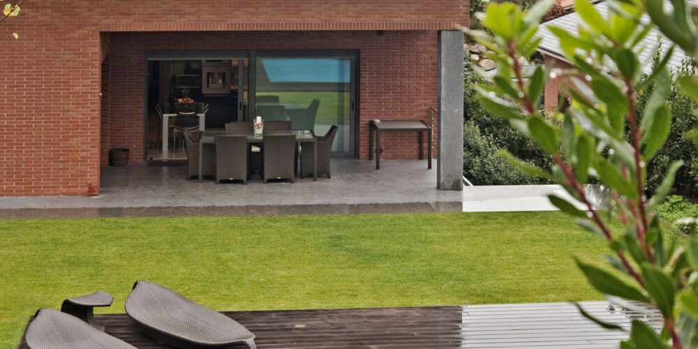 Un entorno al aire libre moderno que muestra una casa de ladrillo contemporánea con grandes puertas de vidrio, que conducen a un patio bellamente amueblado, ideal para comer al aire libre. La escena incluye sillas de lounge negras y elegantes en una terraza de madera, una piscina con rayas azules y un exuberante césped verde rodeado de árboles y arbustos.