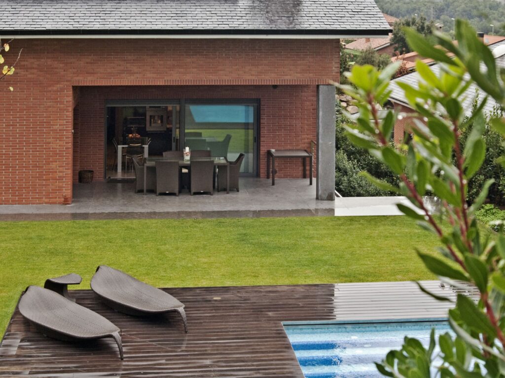 Espacio Exterior Elegante con una Casa de Ladrillo Contemporánea y Piscina Un entorno al aire libre moderno que muestra una casa de ladrillo contemporánea con grandes puertas de vidrio, que conducen a un patio bellamente amueblado, ideal para comer al aire libre. La escena incluye sillas de lounge negras y elegantes en una terraza de madera, una piscina con rayas azules y un exuberante césped verde rodeado de árboles y arbustos.