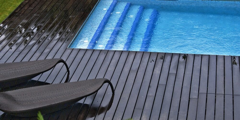Una piscina rectangular ubicada en un entorno al aire libre sereno, adornada con suelos de madera oscura. Sillas modernas de descanso están colocadas cerca, y la piscina refleja el cielo arriba. La vegetación frondosa y las plantas en flor rodean el área, mejorando la atmósfera tranquila con algunas gotas de lluvia visibles en la plataforma de madera por una reciente lluvia.