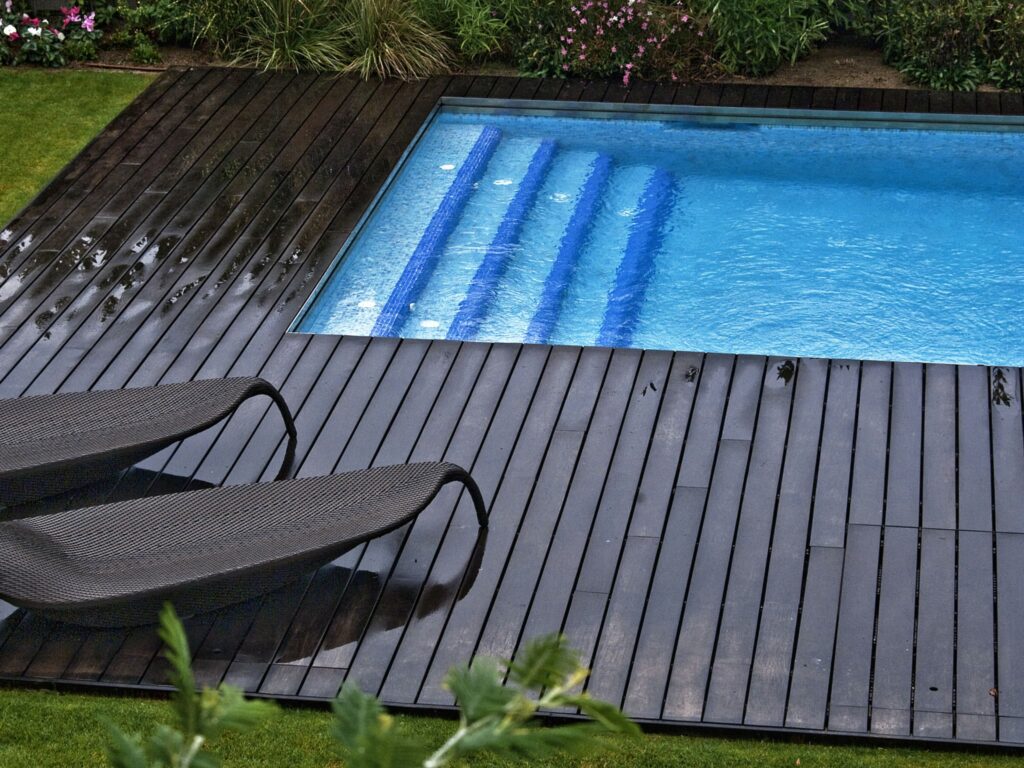 Oasis de Piscina Exterior Tranquila con Vegetación Frondosa Una piscina rectangular ubicada en un entorno al aire libre sereno, adornada con suelos de madera oscura. Sillas modernas de descanso están colocadas cerca, y la piscina refleja el cielo arriba. La vegetación frondosa y las plantas en flor rodean el área, mejorando la atmósfera tranquila con algunas gotas de lluvia visibles en la plataforma de madera por una reciente lluvia.