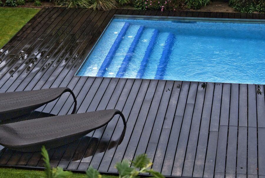 Una piscina rectangular ubicada en un entorno al aire libre sereno, adornada con suelos de madera oscura. Sillas modernas de descanso están colocadas cerca, y la piscina refleja el cielo arriba. La vegetación frondosa y las plantas en flor rodean el área, mejorando la atmósfera tranquila con algunas gotas de lluvia visibles en la plataforma de madera por una reciente lluvia.