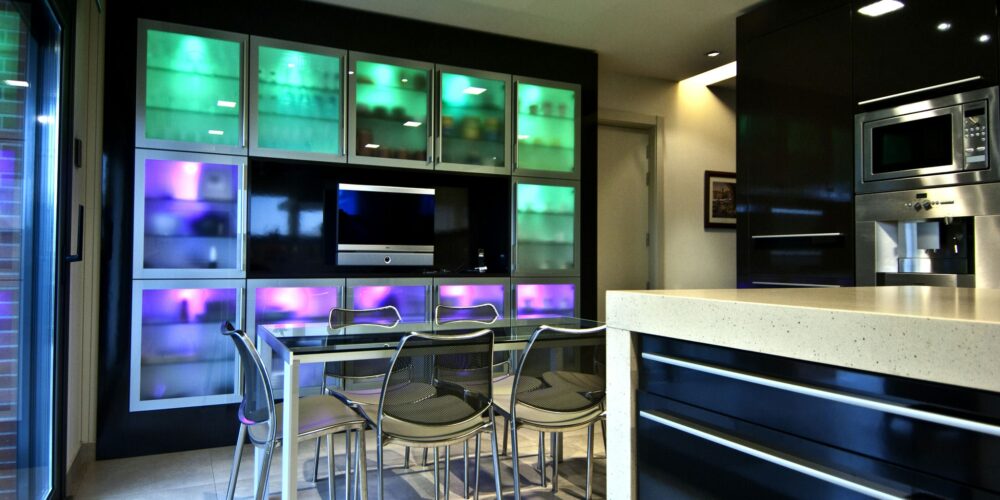 Una cocina moderna con un diseño elegante y un gabinete de exhibición iluminado en luces LED verdes y moradas, que muestra varios elementos. La cocina incluye una encimera de color claro, gabinetes oscuros y sillas de metal contemporáneo alrededor de una mesa de comedor de vidrio, con grandes puertas de vidrio que conducen a un espacio exterior.