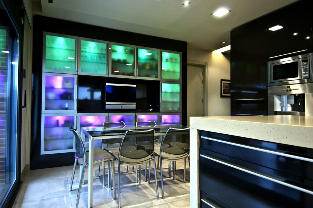 Moderna Cocina Interior Elegante con Gabinete de Exhibición Iluminado por LED Una cocina moderna con un diseño elegante y un gabinete de exhibición iluminado en luces LED verdes y moradas, que muestra varios elementos. La cocina incluye una encimera de color claro, gabinetes oscuros y sillas de metal contemporáneo alrededor de una mesa de comedor de vidrio, con grandes puertas de vidrio que conducen a un espacio exterior.