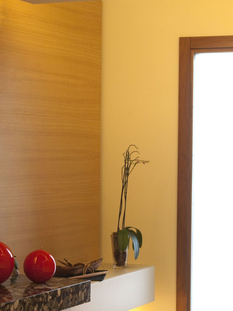 Interior Minimalista Contemporáneo con Esferas Decorativas Rojas y Planta de Orquídea Un interior moderno y elegante que presenta una estantería minimalista con esferas decorativas rojas brillantes, un par de gafas y una planta de orquídea esbelta en un recipiente de vidrio, contra un fondo de madera clara iluminado por una suave luz natural proveniente de una gran ventana.