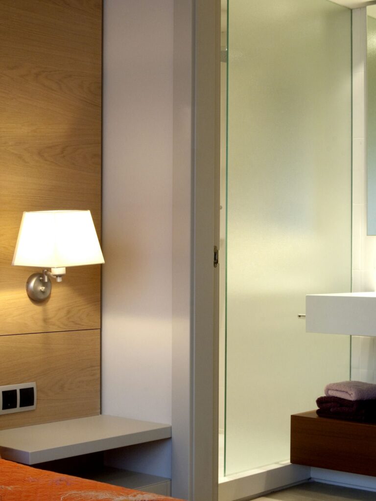 Habitación de hotel moderna y acogedora con decoración de madera y elegante baño Una esquina estilosa de una habitación de hotel que presenta paneles de madera cálida, una mesa de noche minimalista con una lámpara y un panel de vidrio esmerilado que separa el área de descanso de un elegante baño con toallas organizadas de manera ordenada.