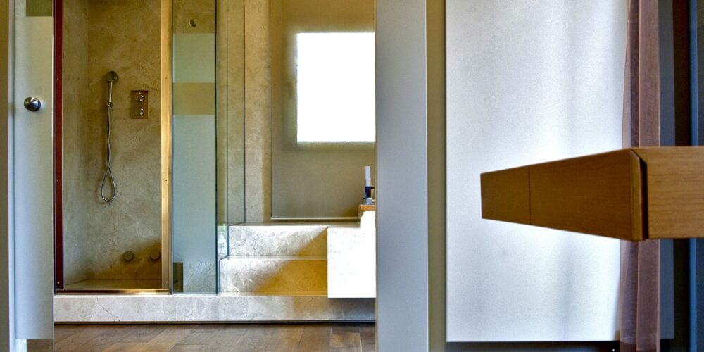 Un interior de baño contemporáneo que muestra una ducha cerrada de cristal a la izquierda con grifos elegantes y paredes de piedra de color claro, iluminado por la luz del sol que entra a través de una gran ventana. El suelo de madera oscura contrasta con los tonos claros, mientras que a la derecha se ve un tocador de madera funcional.