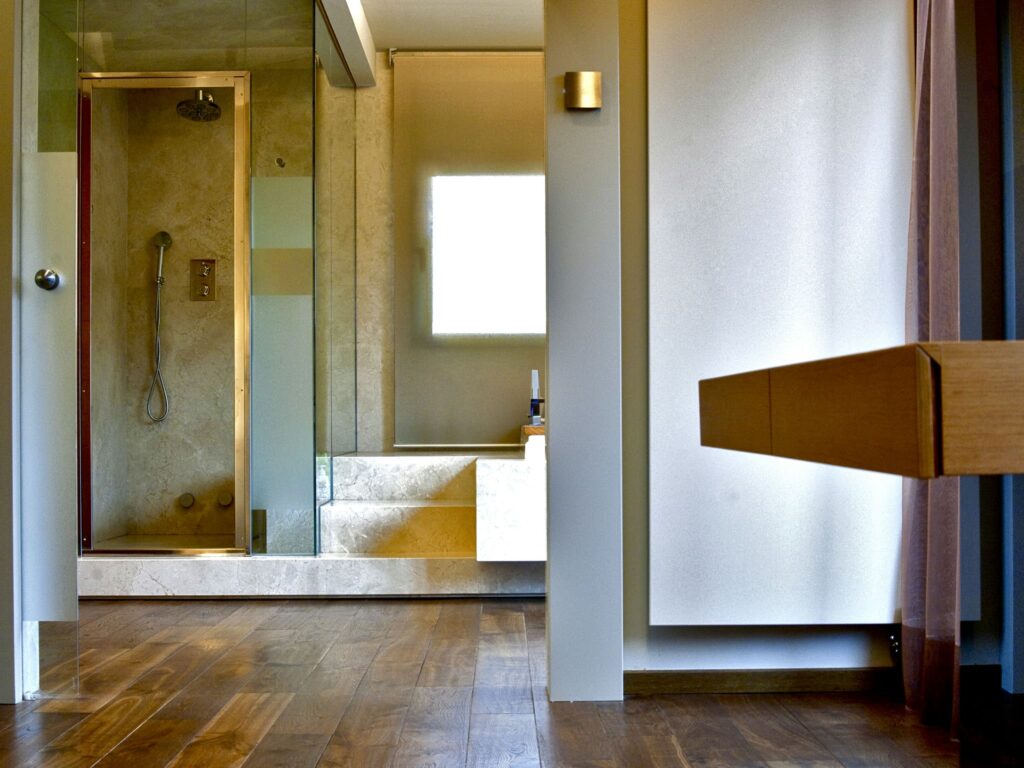 Baño Moderno Minimalista con Ducha Cerrada de Cristal y Luz Natural Un interior de baño contemporáneo que muestra una ducha cerrada de cristal a la izquierda con grifos elegantes y paredes de piedra de color claro, iluminado por la luz del sol que entra a través de una gran ventana. El suelo de madera oscura contrasta con los tonos claros, mientras que a la derecha se ve un tocador de madera funcional.