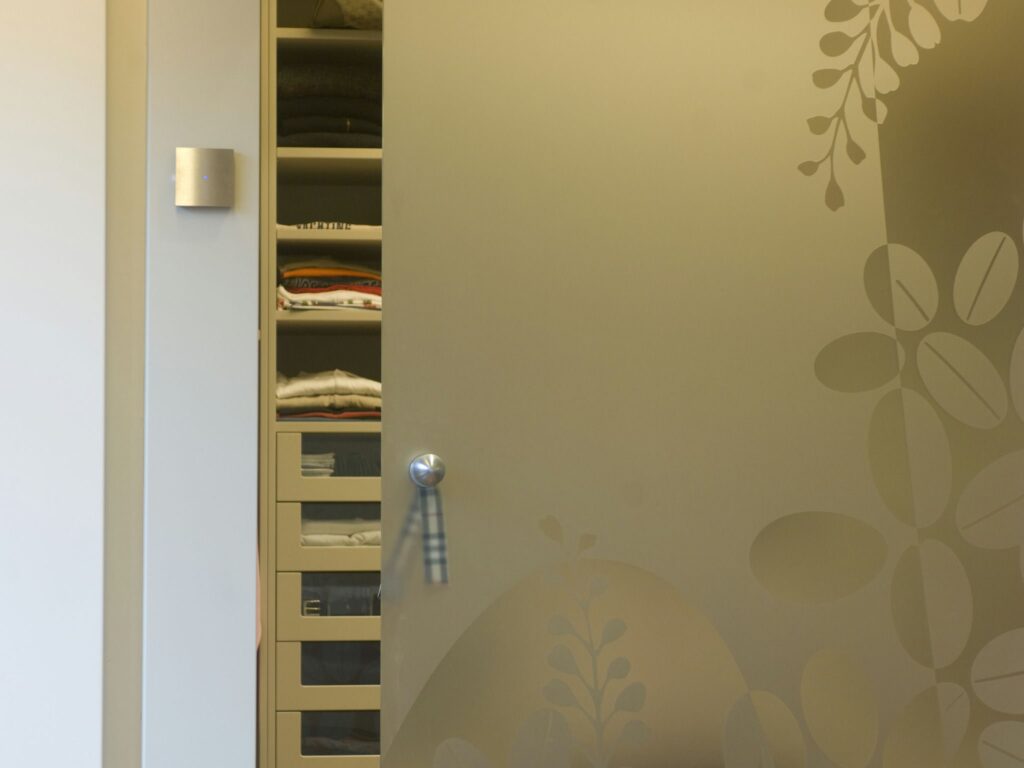 Armario Minimalista Moderno con Almacenamiento de Ropa Organizado Una puerta de armario moderno parcialmente abierta que revela artículos de ropa dispuestos ordenadamente y cajones de almacenamiento, con un diseño minimalista que incluye vidrio esmerilado y sutiles patrones de hojas, ubicado en un espacio bien iluminado que enfatiza el orden y el estilo contemporáneo.
