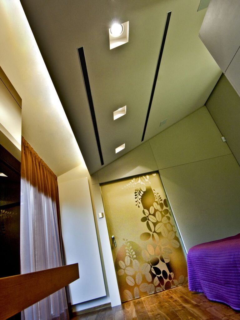 Interiores Modernos Elegantes con Techo Único y Puerta de Vidrio Decorativa Un interior de habitación elegante que presenta un techo único con iluminación empotrada, suelos de madera y una gran puerta de vidrio decorativa con intrincados patrones de hojas. La decoración incluye un cubrecamas morado y cortinas suaves, lo que contribuye a una atmósfera contemporánea y acogedora.