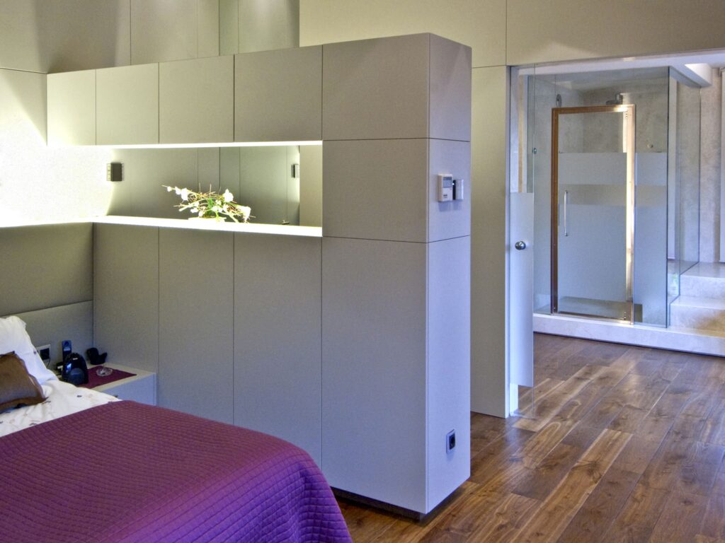 Interior de Dormitorio Moderno con Elementos de Diseño Contemporáneo Un sofisticado dormitorio moderno que presenta una cama grande adornada con una rica manta púrpura, cabinetry de color claro elegante con iluminación incorporada y una cabina de ducha de vidrio transparente, todo sobre un cálido suelo de madera pulida, que encarna el minimalismo y la comodidad.