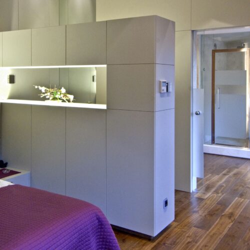Un sofisticado dormitorio moderno que presenta una cama grande adornada con una rica manta púrpura, cabinetry de color claro elegante con iluminación incorporada y una cabina de ducha de vidrio transparente, todo sobre un cálido suelo de madera pulida, que encarna el minimalismo y la comodidad.