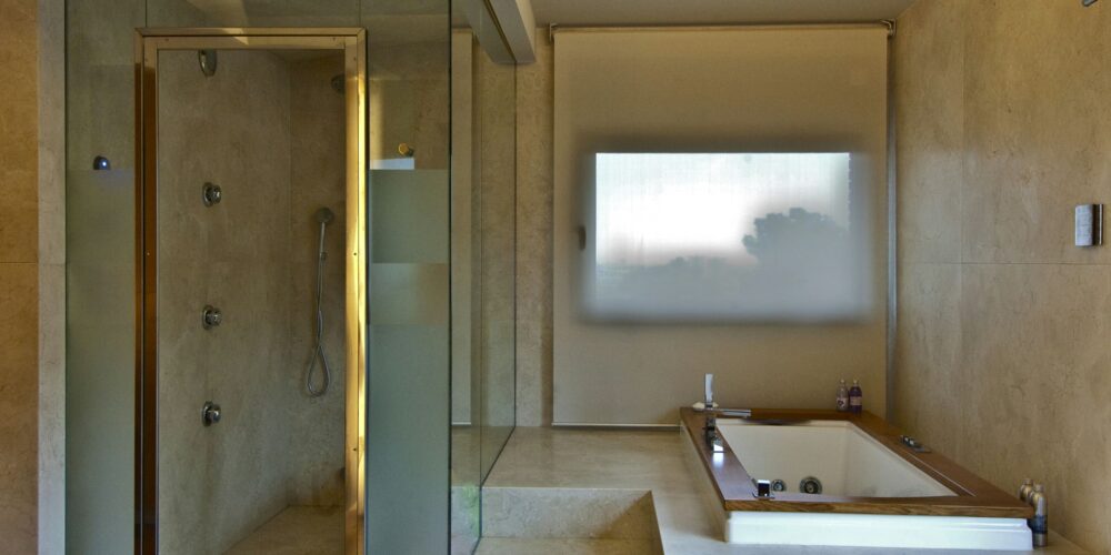 Un baño contemporáneo que presenta una moderna ducha cerrada en vidrio con múltiples cabezales de ducha, una espaciosa bañera de hidromasaje en una plataforma elevada, un suelo de azulejos de piedra de color claro y una ventana esmerilada para luz natural.