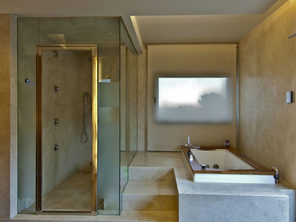 Baño Moderno de Lujo con Ducha Cerrada en Vidrio y Bañera de Hidromasaje Un baño contemporáneo que presenta una moderna ducha cerrada en vidrio con múltiples cabezales de ducha, una espaciosa bañera de hidromasaje en una plataforma elevada, un suelo de azulejos de piedra de color claro y una ventana esmerilada para luz natural.