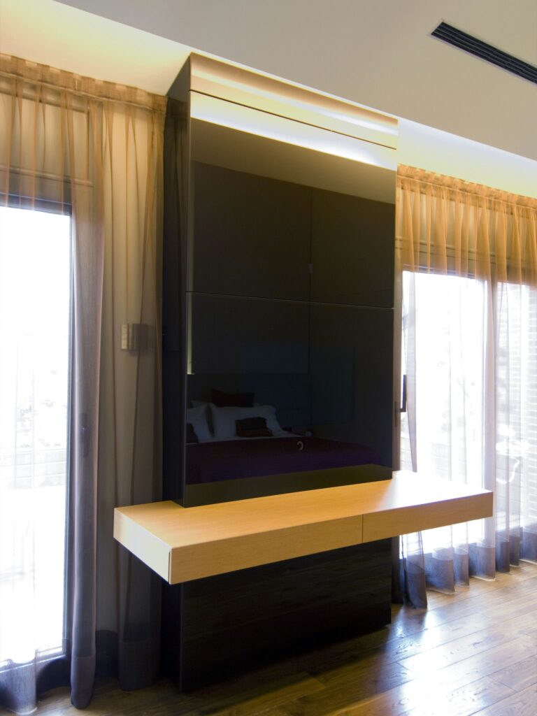 Pieza de Mobiliario Moderno Elegante con Diseño Sleek y Acabado Brillante Una instalación de muebles moderna y elegante que presenta un panel oscuro y brillante que sirve como unidad montada en la pared y estante flotante, complementada por una superficie de madera clara ideal para trabajar o exhibir, con cortinas ligeras en el fondo filtrando la luz natural.