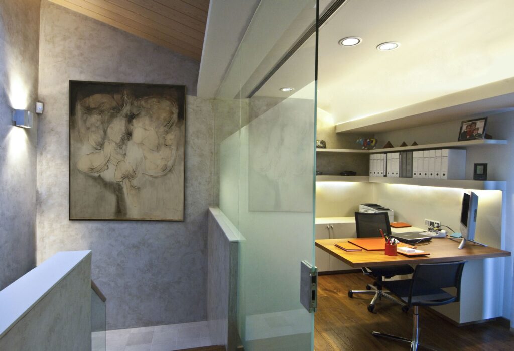 Espacio de oficina moderno con partición de vidrio y diseño contemporáneo Un interior de oficina moderna que presenta una partición de vidrio que separa efectivamente el espacio de trabajo del pasillo. La imagen muestra un elegante escritorio de madera acompañado de una silla ergonómica, una configuración de computadora y suministros organizados de manera ordenada. Las paredes están decoradas con una vibrante pintura abstracta y estanterías que albergan varios artículos y archivos. Una suave iluminación ambiental mejora la estética contemporánea, complementada por un cálido suelo de madera, creando un ambiente acogedor y productivo.