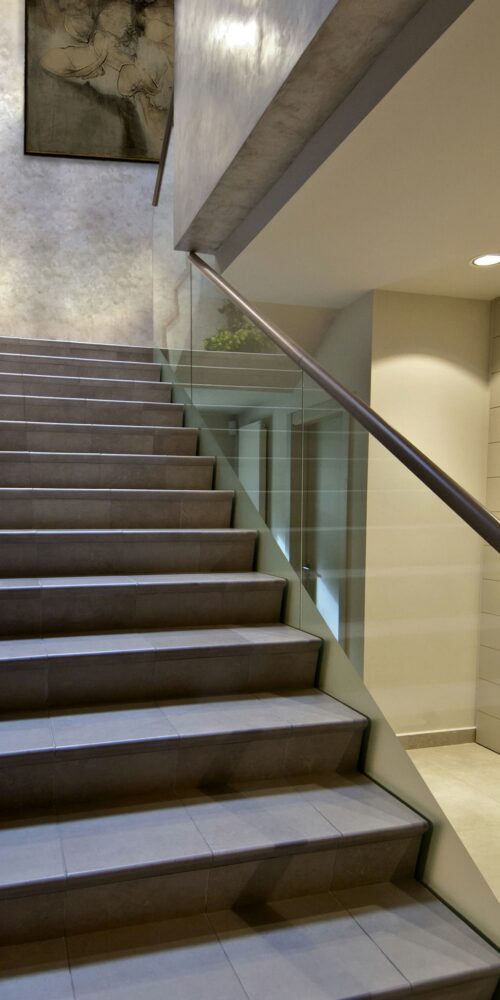 Una escalera moderna con suaves baldosas grises y una elegante barandilla de vidrio, iluminada por una suave luz. Por encima del descanso, una obra de arte abstracto realza la decoración minimalista, mostrando líneas limpias y un diseño contemporáneo.