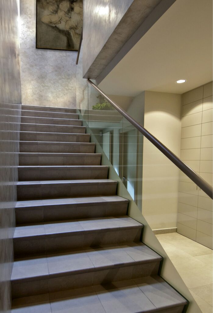 Escalera interior contemporánea con barandilla de vidrio y arte abstracto Una escalera moderna con suaves baldosas grises y una elegante barandilla de vidrio, iluminada por una suave luz. Por encima del descanso, una obra de arte abstracto realza la decoración minimalista, mostrando líneas limpias y un diseño contemporáneo.