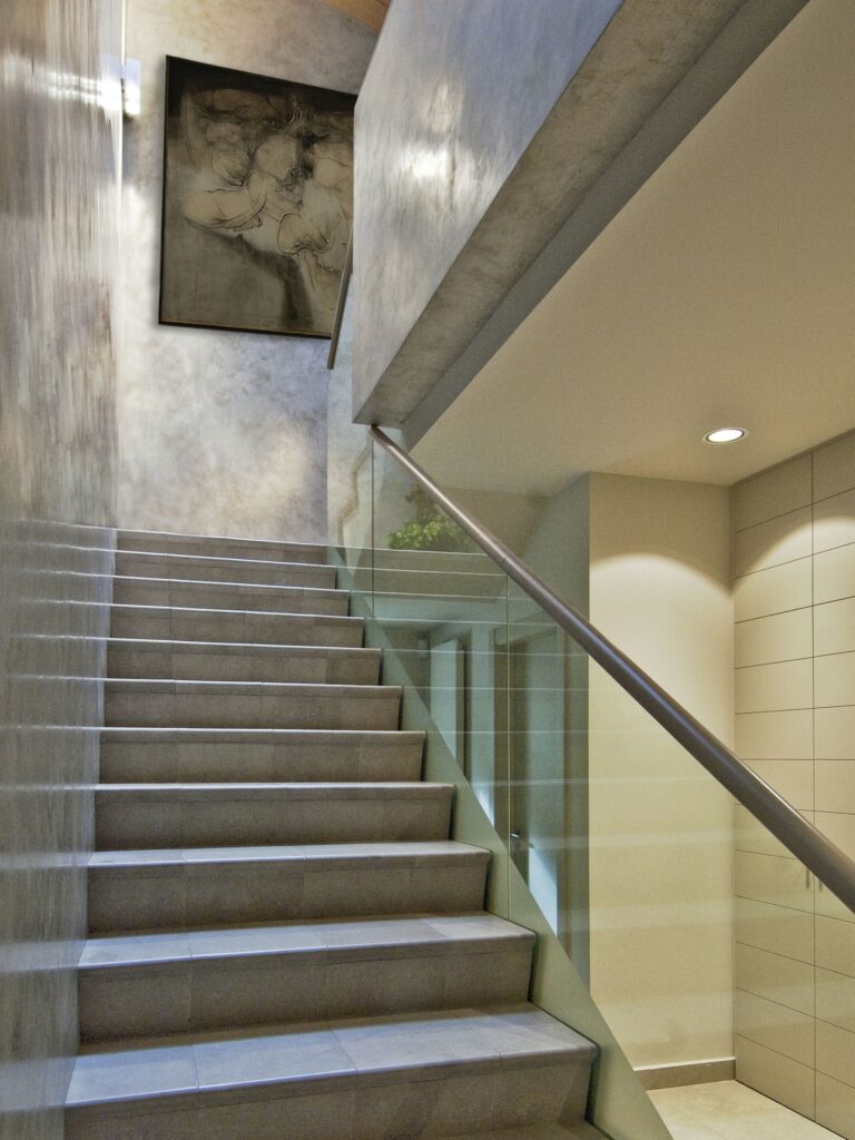 Escalera moderna con peldaños de cerámica gris y barandilla de vidrio Una escalera contemporánea que presenta peldaños de cerámica gris y una elegante barandilla de vidrio, iluminada por una suave iluminación. Sobre la escalera hay una pared texturizada con una gran pintura abstracta, que contribuye a un diseño interior elegante y minimalista.